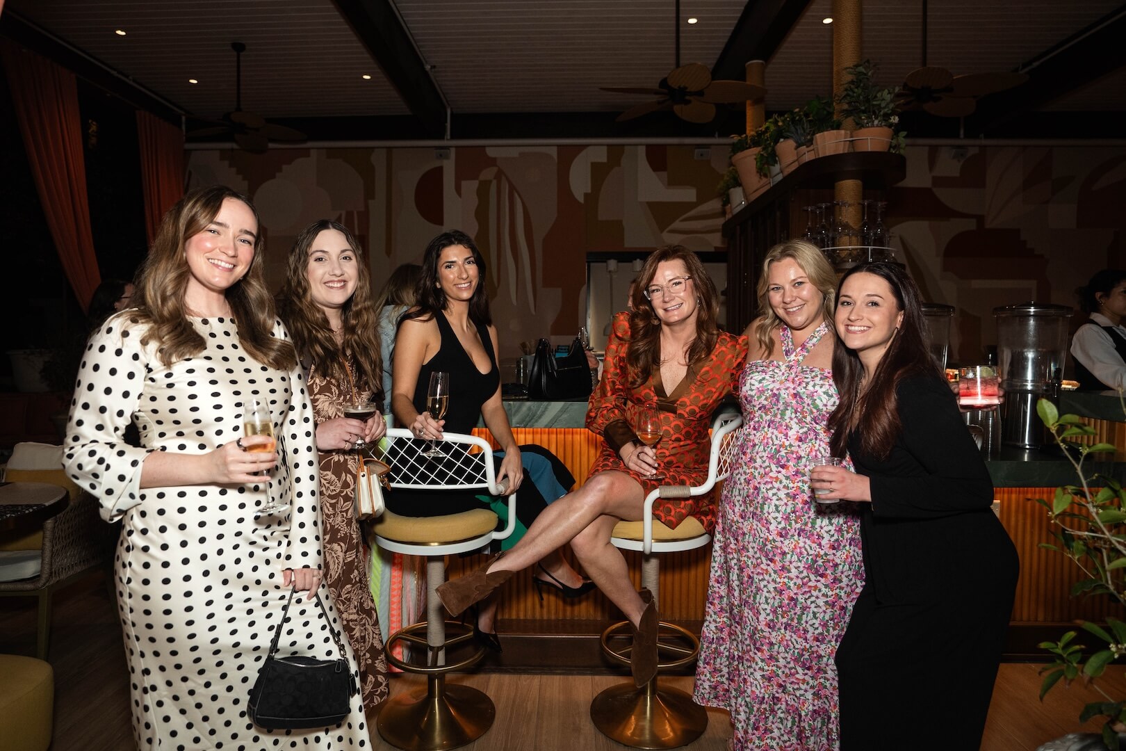 Six women in dresses pose and smile for a group photo at an indoor social gathering, with drinks in hand and a bar in the background. - HC Beauty in Charlotte, Nc