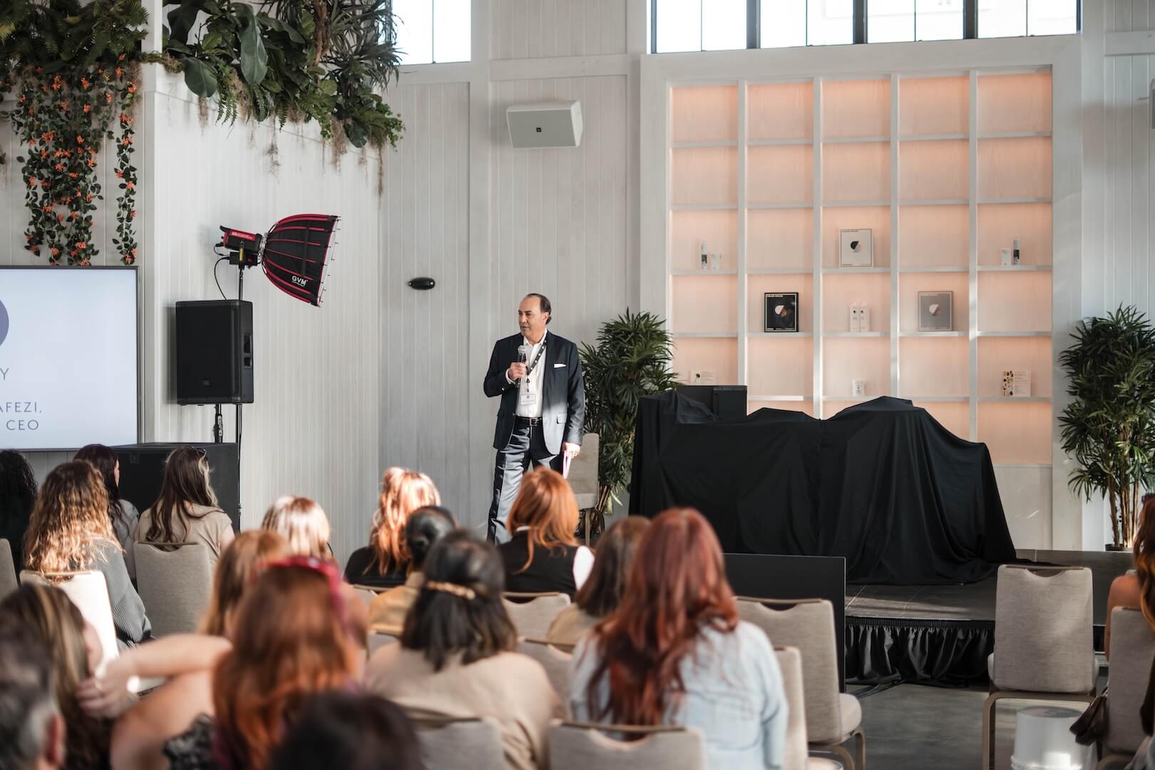 A man in a suit speaks on stage to a seated audience in a modern, well-lit event space with plants and shelving in the background. - HC Beauty in Charlotte, Nc