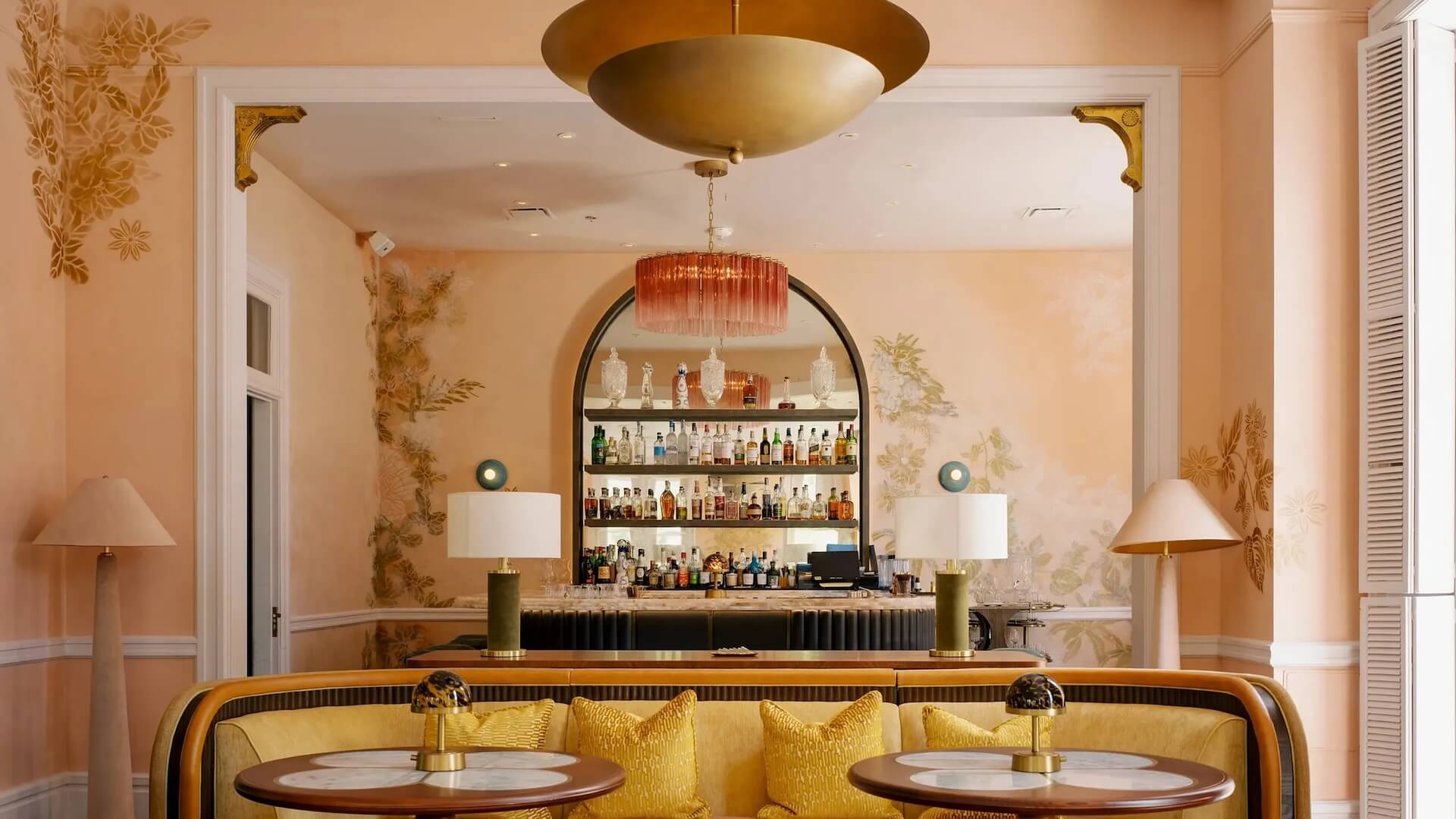 A stylish bar area with peach walls, a large arched mirror, shelves of liquor bottles, gold accents, a chandelier, and a yellow sofa with round tables. - HC Beauty in Charlotte, Nc